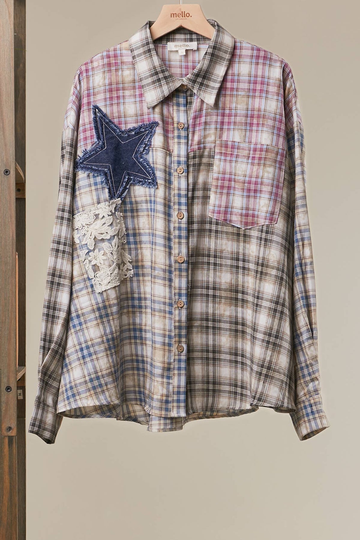 A long-sleeve plaid button-up shirt hanging on a wooden hanger, featuring a blue denim star patch and a lace applique pocket on the left side. The shirt has a beige, blue, and pink plaid pattern with brown buttons.