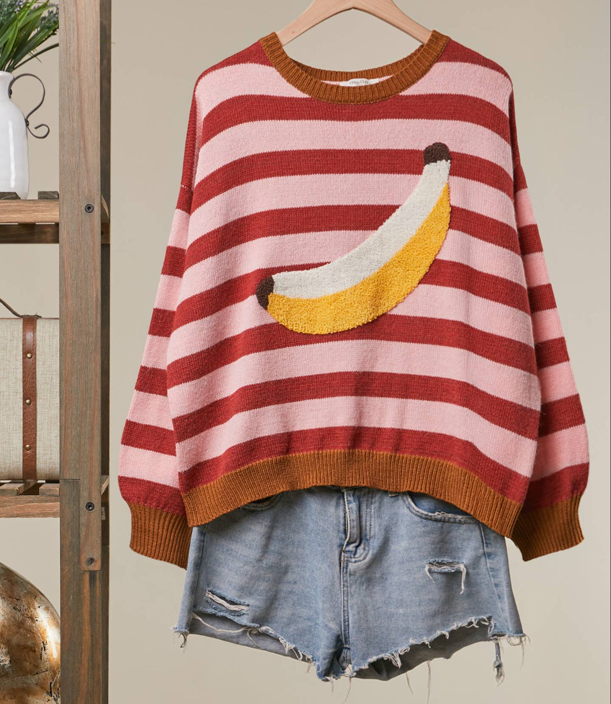 A pink and red striped knit sweater with a yellow banana applique on the front, hanging on a wooden hanger with light-wash denim shorts underneath, against a beige wall with a wooden shelf on the left.