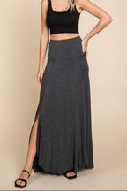 A woman modeling a dark gray knit maxi skirt with a side slit, paired with a black crop top and black sandals, against a solid beige studio background.