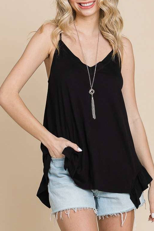 A smiling blonde woman wearing a black sleeveless camisole top with a ruffled hem and light blue denim shorts, standing against a plain beige studio background.