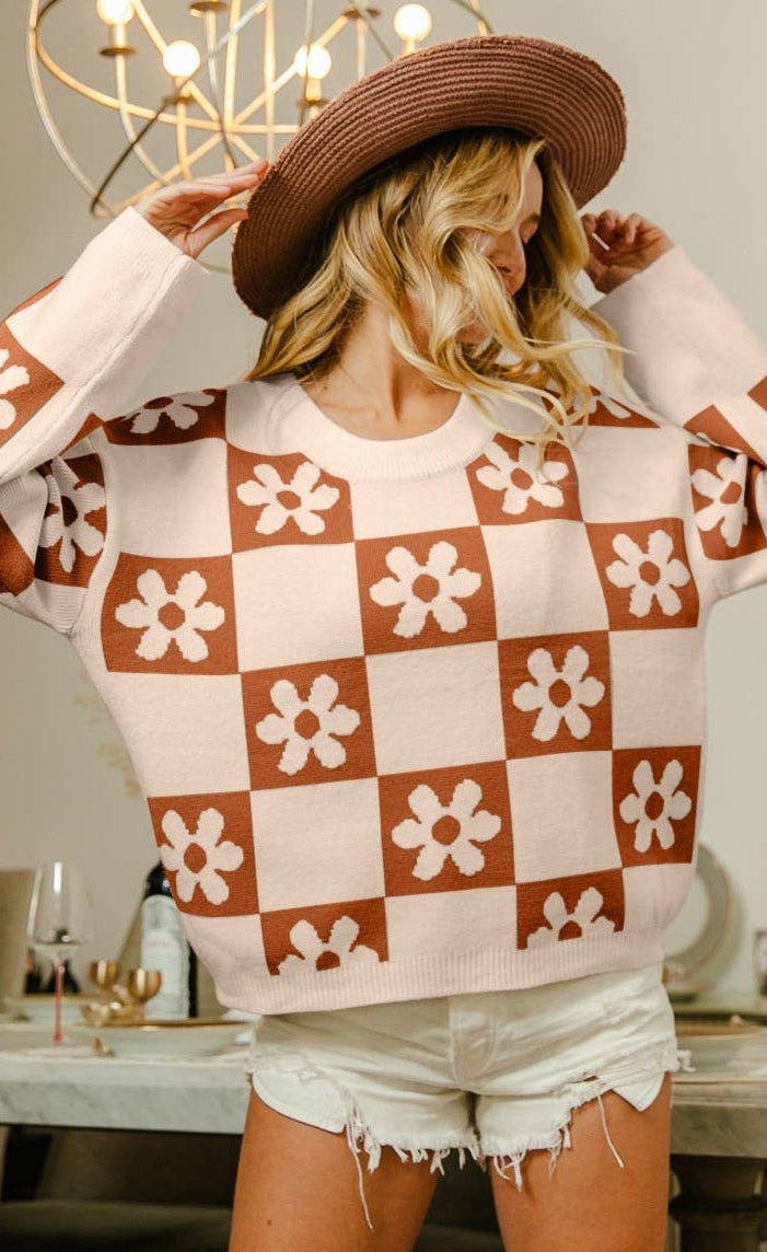 A woman wearing a brown and cream checkerboard knit sweater with white flower motifs, a brown wide-brim hat, and white denim shorts, standing in a dining room setting.