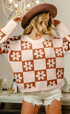 A woman wearing a brown and cream checkerboard knit sweater with white flower motifs, a brown wide-brim hat, and white denim shorts, standing in a dining room setting.