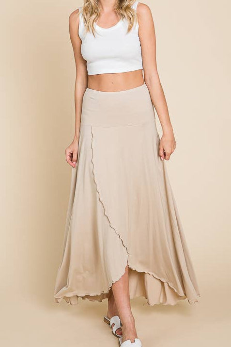 A woman modeling a beige wrap-style midi skirt with a ruffled hem, paired with a white crop top, wide-brim straw hat, and white slide sandals, against a solid beige studio background.