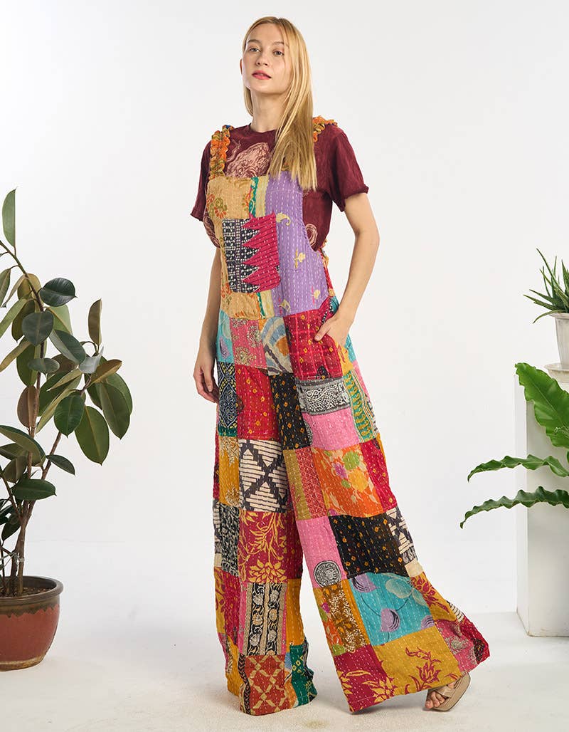 Woman wearing a colorful patchwork dress standing between two potted plants on a white background