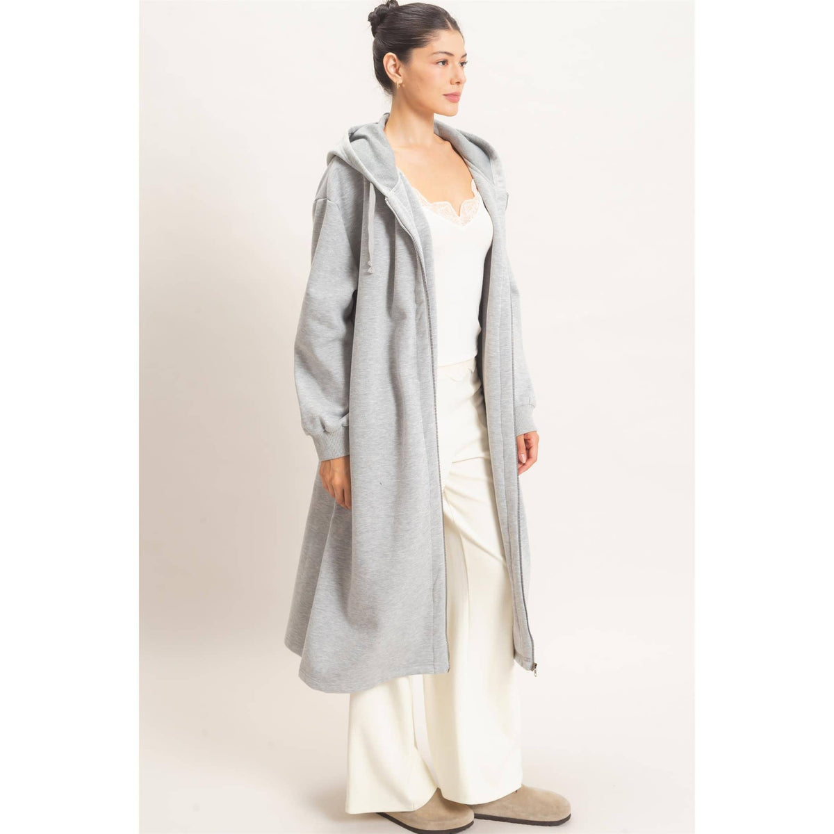 A woman modeling a light gray hooded longline cardigan over a white camisole and white wide-leg pants, standing against a plain off-white studio background.