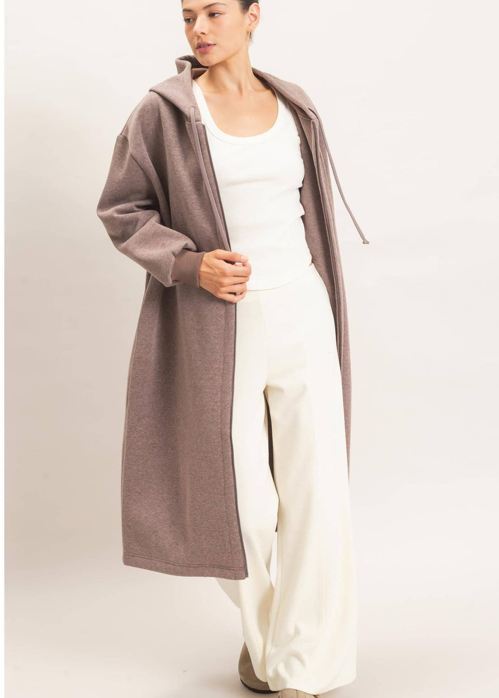 A woman modeling a long, open-front hooded cardigan in a muted taupe-brown color, worn over a white tank top and white wide-leg pants, against a plain light beige studio background.