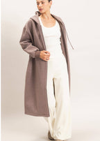 A woman modeling a long, open-front hooded cardigan in a muted taupe-brown color, worn over a white tank top and white wide-leg pants, against a plain light beige studio background.