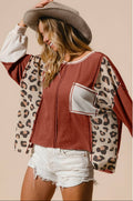 A woman wearing a cropped zip-front top with a rust-brown body, leopard-print sleeves, and a light gray pocket, styled with a wide-brim brown hat and white denim shorts against a plain beige background.