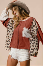 A woman wearing a cropped zip-front top with a rust-brown body, leopard-print sleeves, and a light gray pocket, styled with a wide-brim brown hat and white denim shorts against a plain beige background.