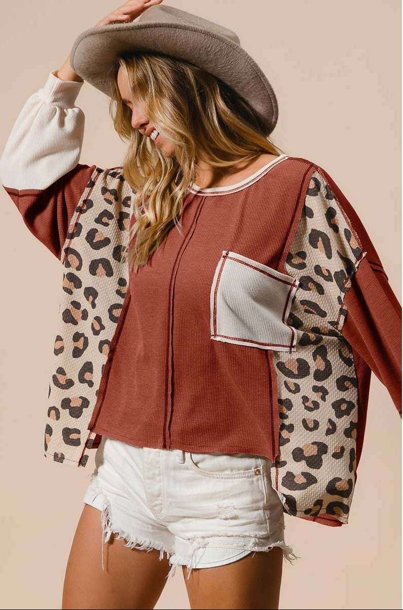 A woman wearing a cropped zip-front top with a rust-brown body, leopard-print sleeves, and a light gray pocket, styled with a wide-brim brown hat and white denim shorts against a plain beige background.