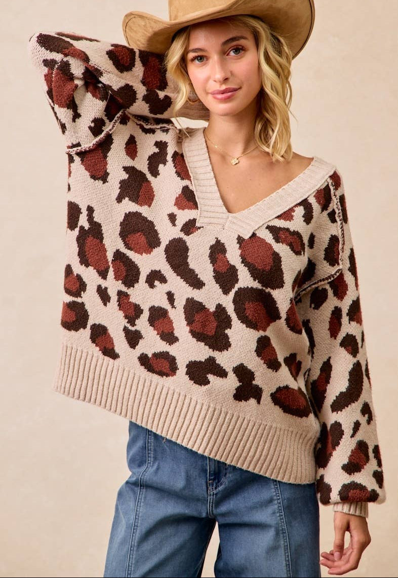 A woman wearing a beige knit sweater with a brown and rust leopard print pattern, paired with blue jeans and a tan cowboy hat, against a solid beige background.