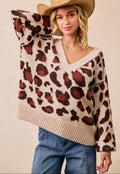 A woman wearing a beige knit sweater with a brown and rust leopard print pattern, paired with blue jeans and a tan cowboy hat, against a solid beige background.