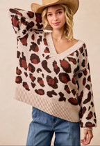 A woman wearing a beige knit sweater with a brown and rust leopard print pattern, paired with blue jeans and a tan cowboy hat, against a solid beige background.