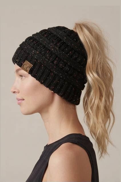 A woman in profile wearing a black ribbed knit ponytail beanie with subtle multicolor flecks and a small tan logo patch on the side, against a plain beige studio background.