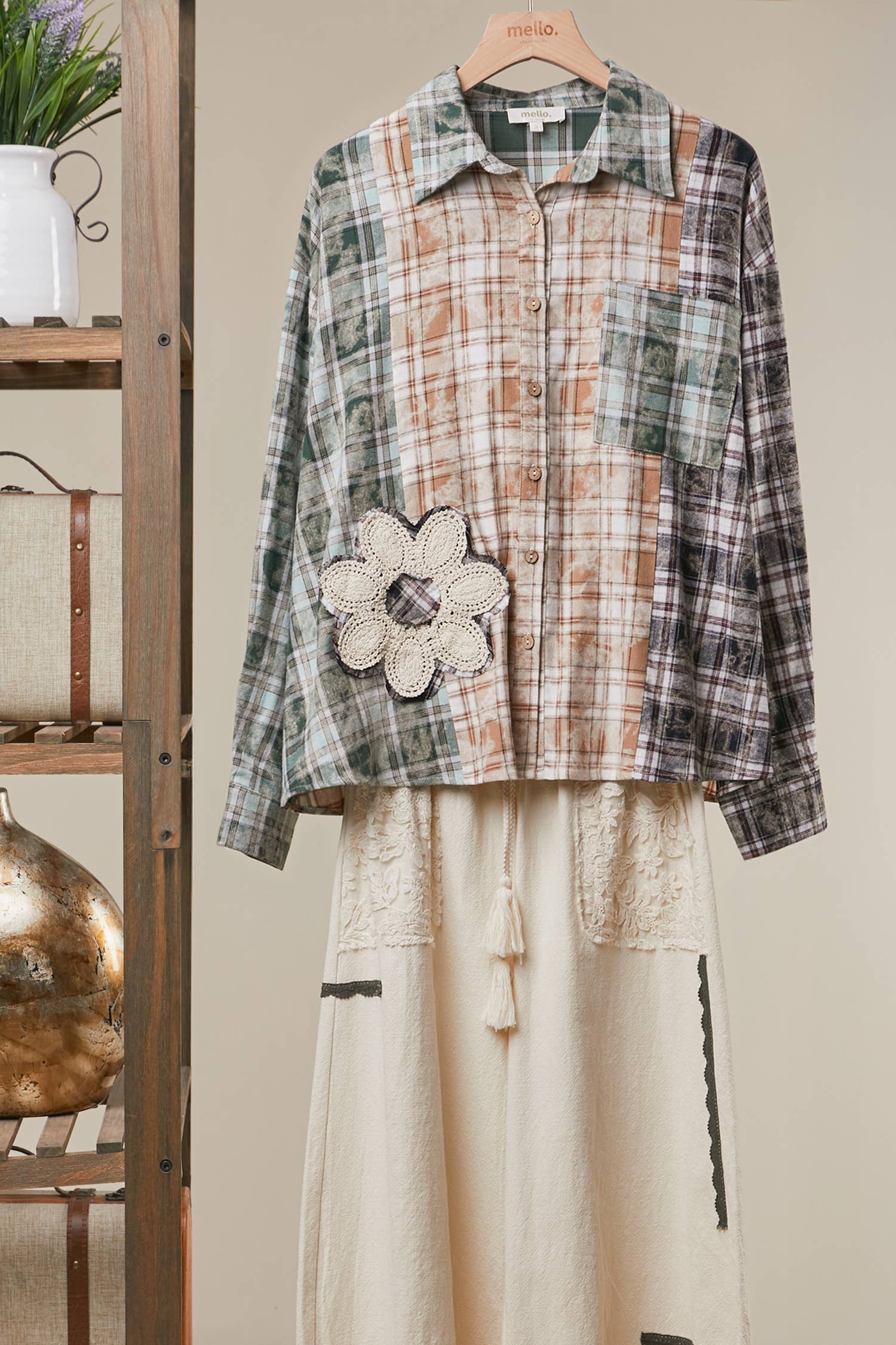 A patchwork plaid shirt jacket with a large crocheted flower applique on the front, paired with a cream-colored skirt, displayed on a wooden hanger against a neutral background.