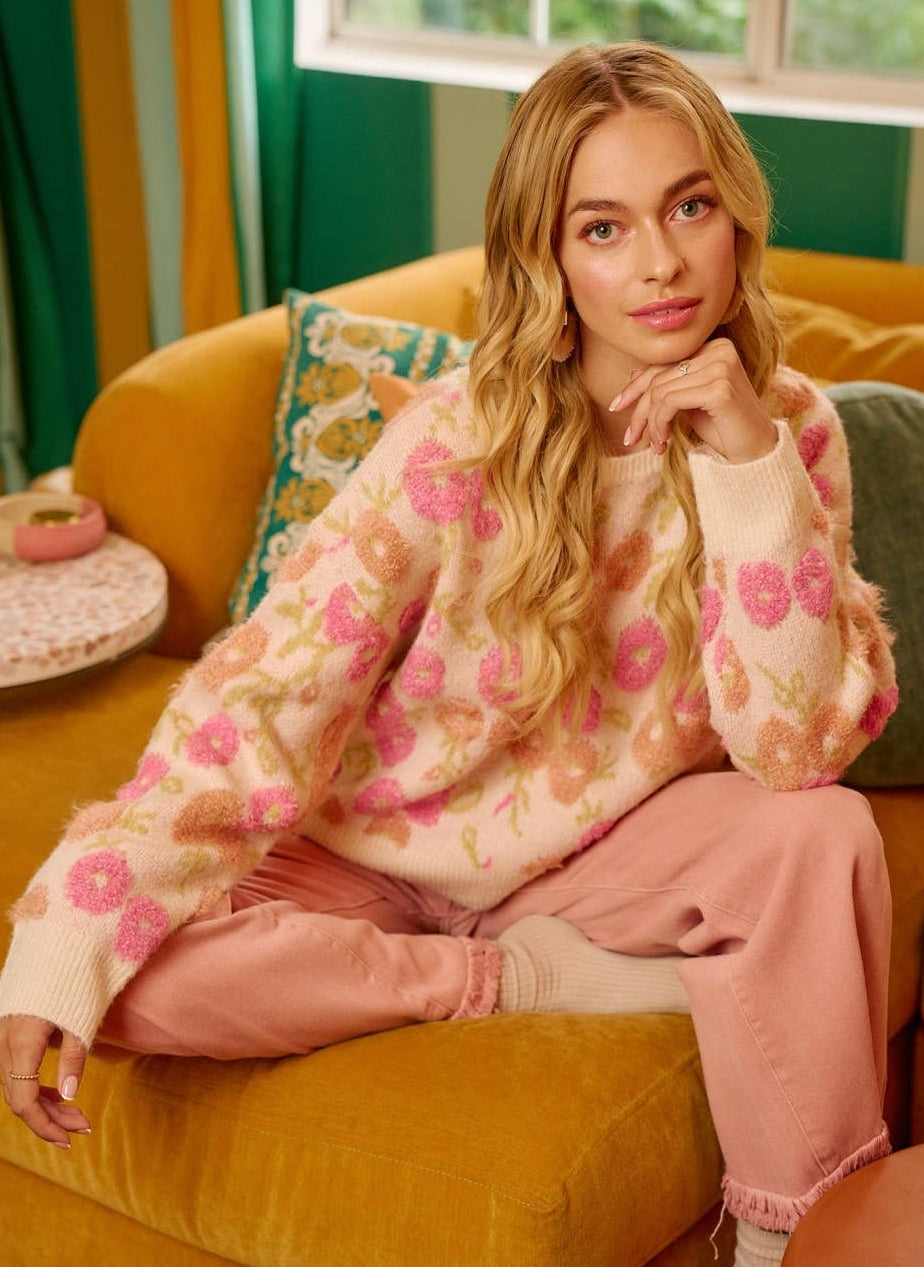 A woman sits on a mustard yellow couch wearing a fuzzy cream knit sweater with pink and peach floral embroidery, paired with pink sweatpants and white socks in a colorful living room setting.