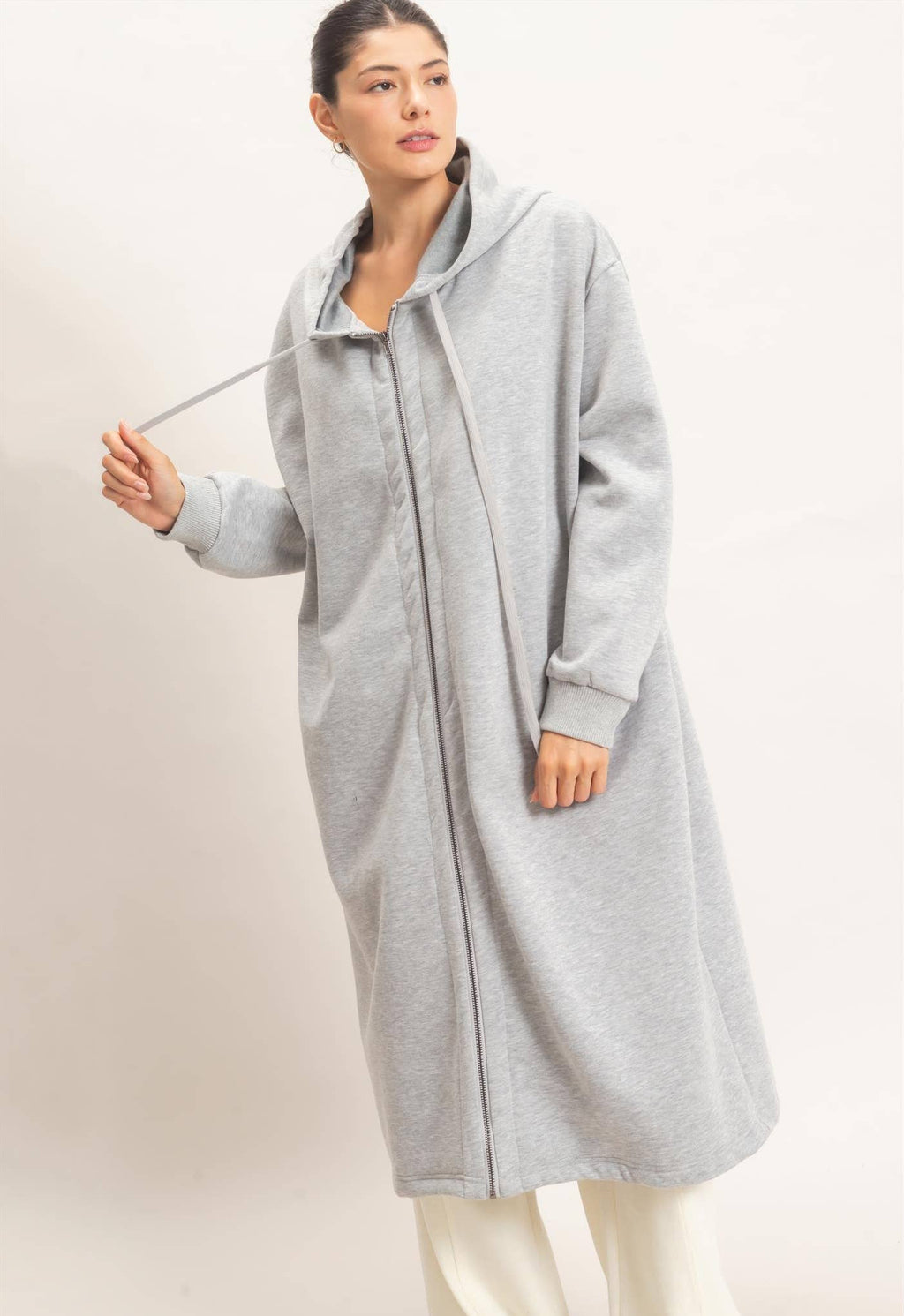 A woman modeling a light gray hooded longline cardigan or coat with a front zipper, shown against a plain light beige studio background.